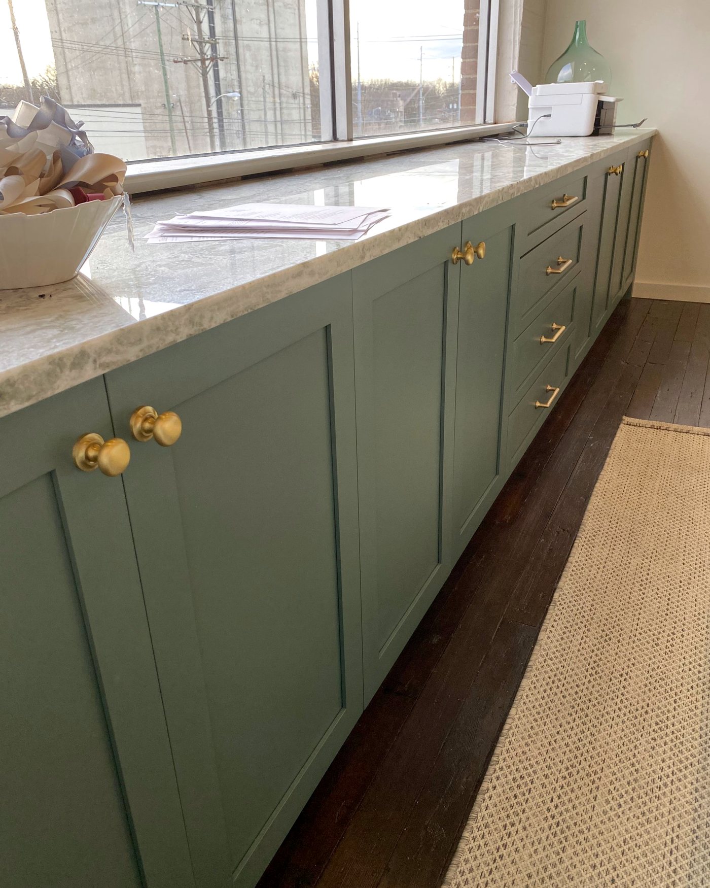 Long run of green painted office cabinetry with brass knobs, a drawer bank and a marble top beneath a wide window