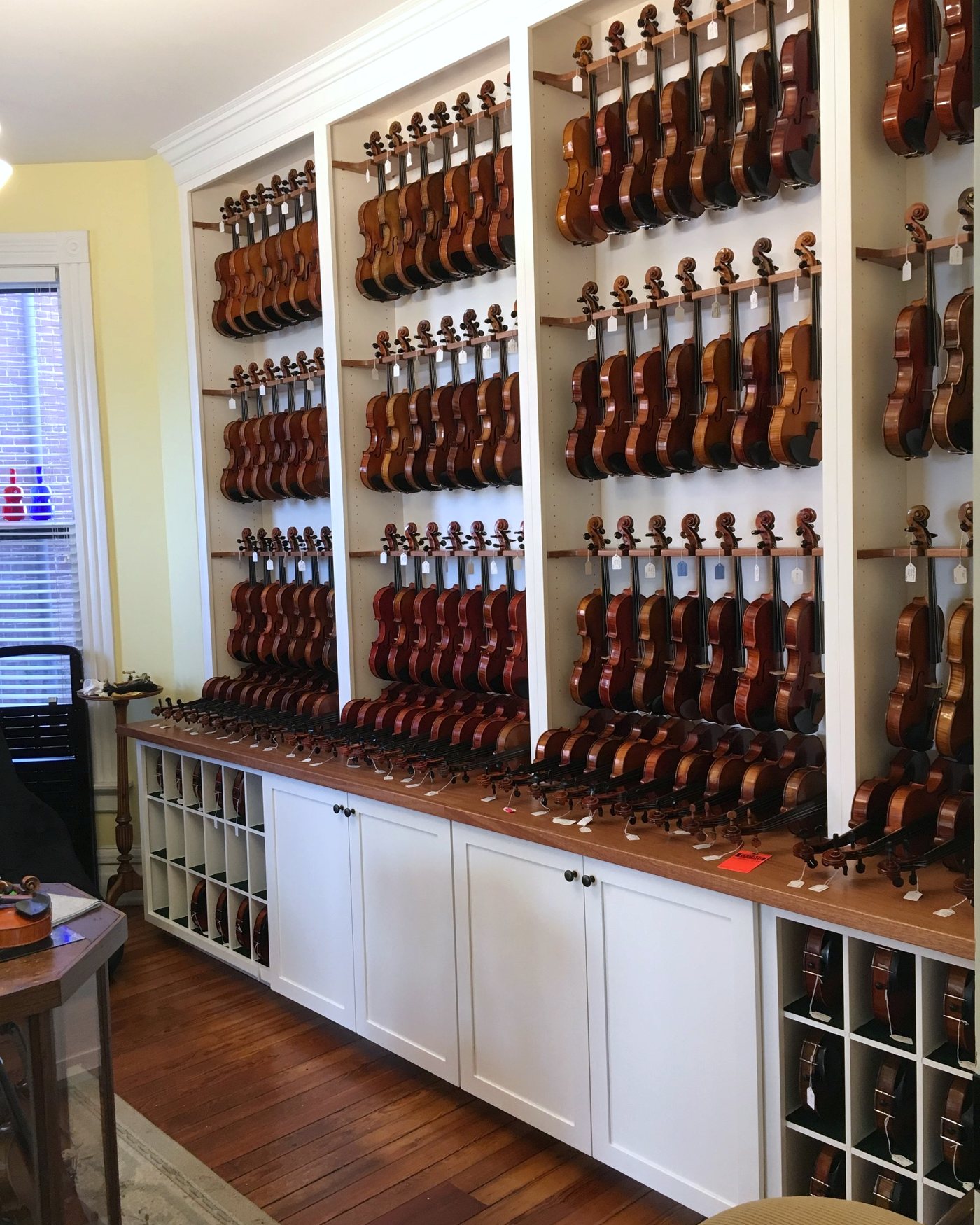 Violin shop display wall: white built-in bays of hanging violins on walnut rails above a counter, cabinets and storage cubbies